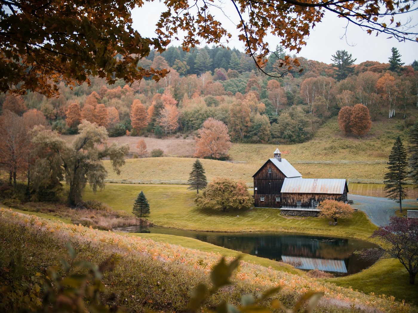 A classic Vermont barn beside a pond, surrounded by rolling hills of autumn foliage